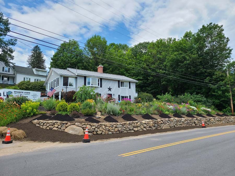 A light-colored house with a stone retaining wall and landscaped flower beds sits beside a road under a partly cloudy sky.