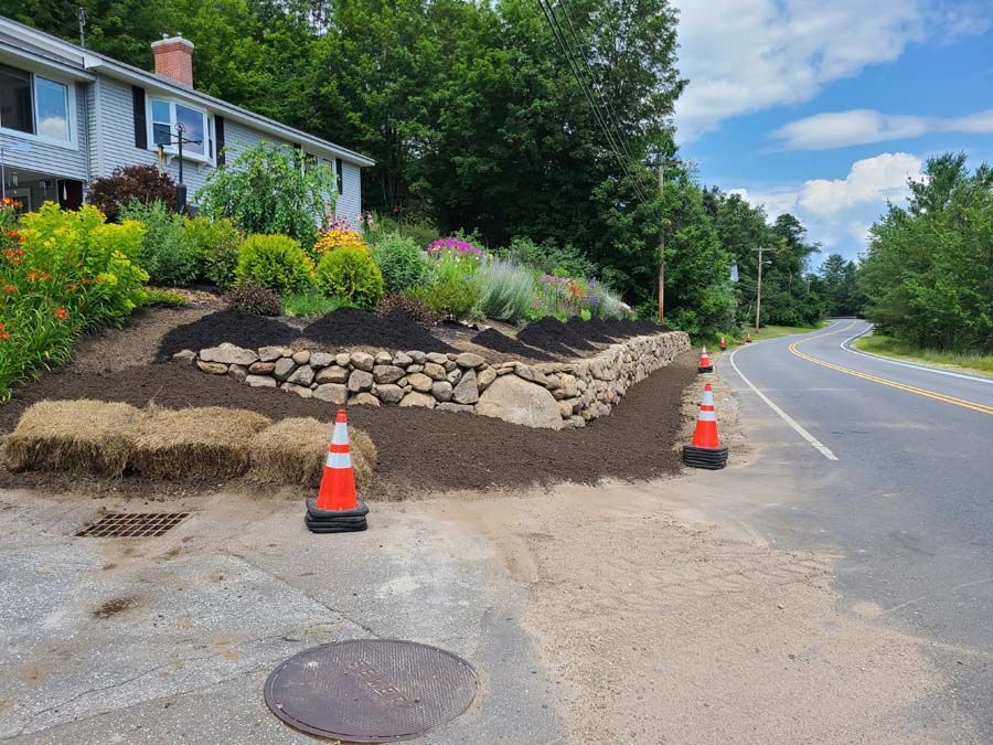 A stone retaining wall built into a landscaped hillside next to a road, featuring fresh mulch, straw bales, and safety cones.