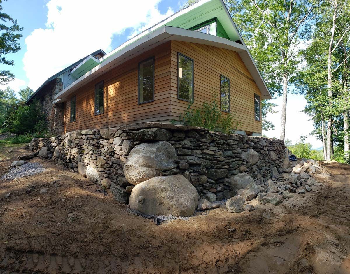 A wooden house sits atop a stone foundation built from large, irregular boulders in a wooded setting under a blue sky.