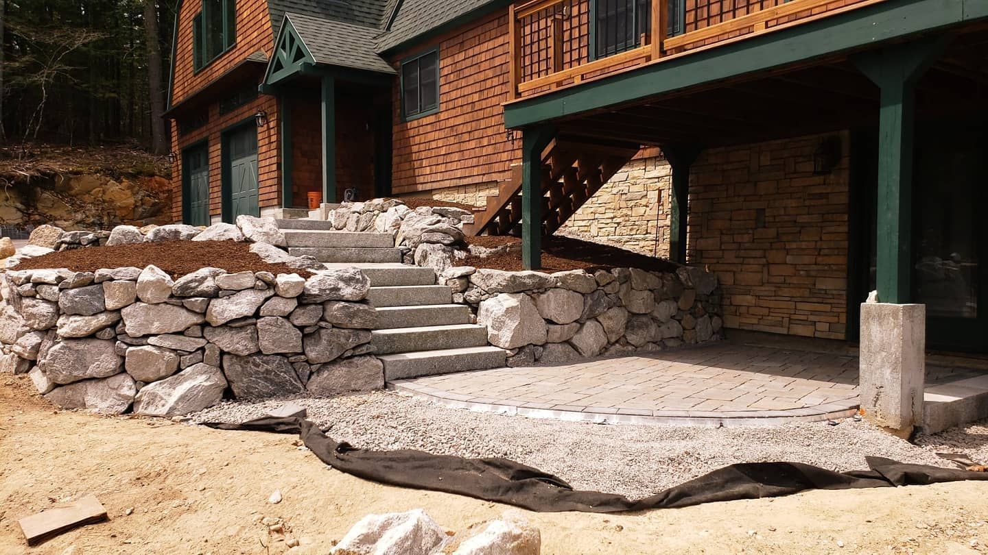 Stone stairs lead up from a gravel patio to a house with a brick exterior and a wooden deck.