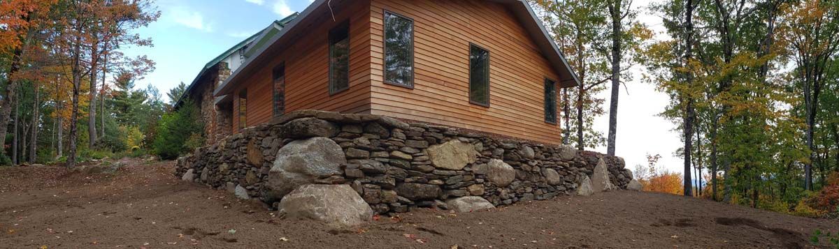 A wooden cabin sits atop a raised stone foundation in a forest during autumn.