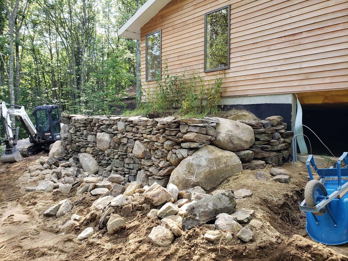 A stone retaining wall built next to a wooden-sided house, with a blue wheelbarrow and construction machinery nearby.