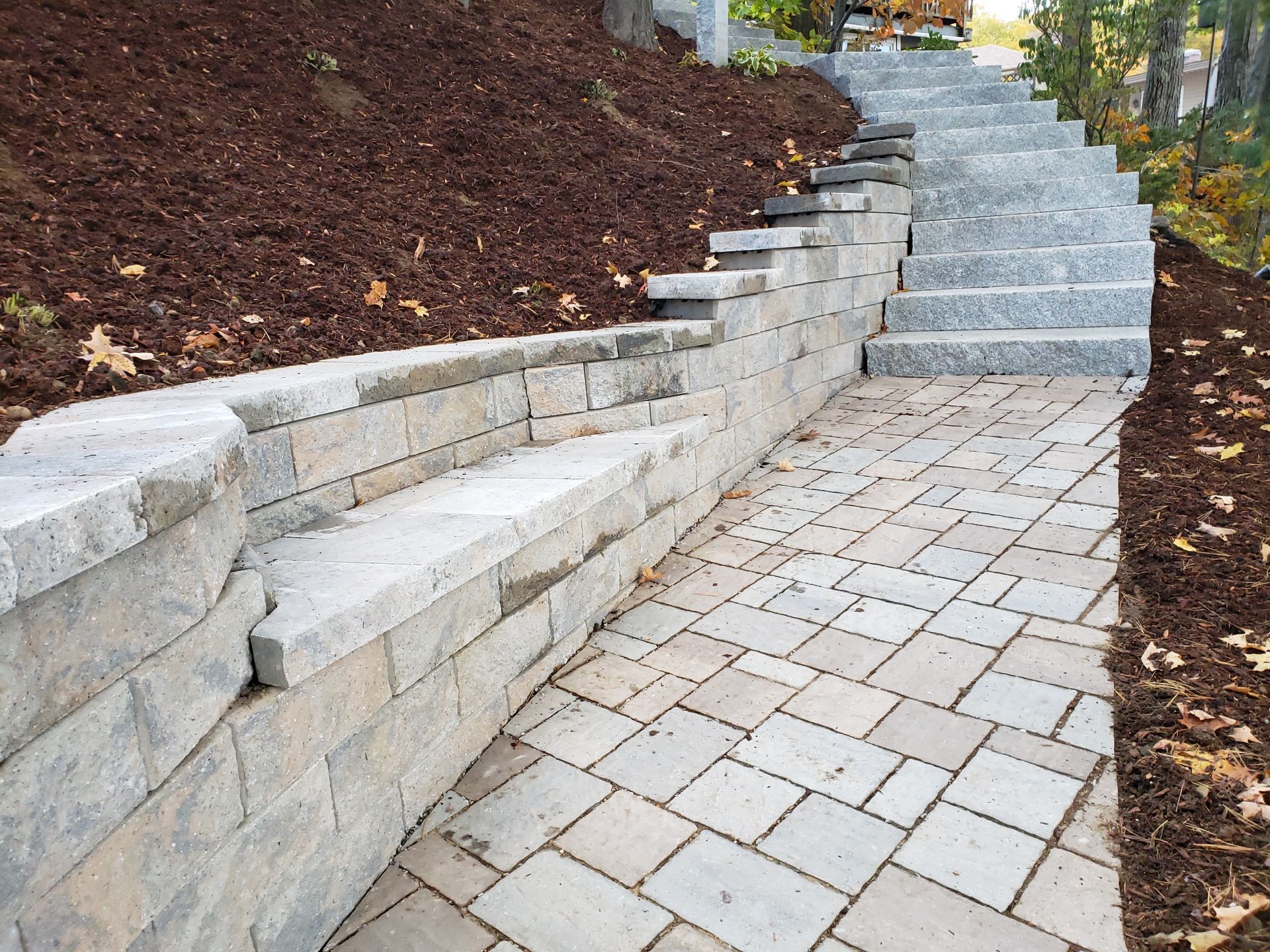 A stone retaining wall with an integrated bench runs alongside a light gray paver path and stone stairs in a landscaped yard.