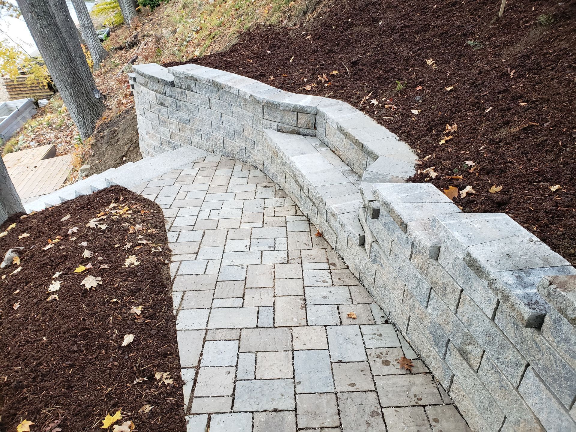 A paved stone walkway curves beside a multi-level retaining wall made of light gray concrete blocks, flanked by mulch.
