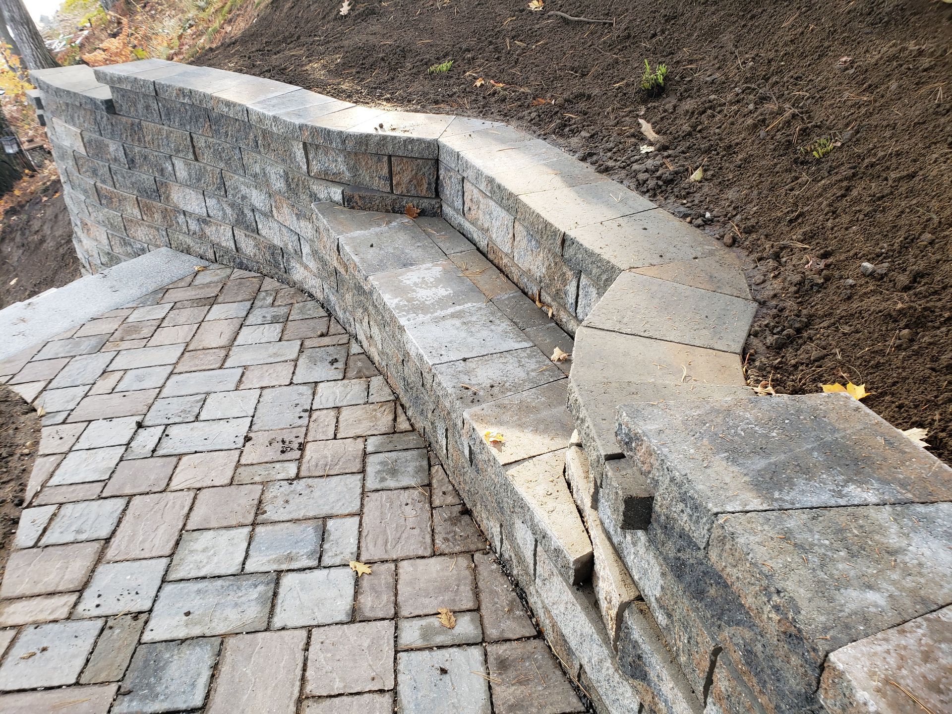A curving stone retaining wall with a built-in bench seat next to a paver patio with soil in the background.