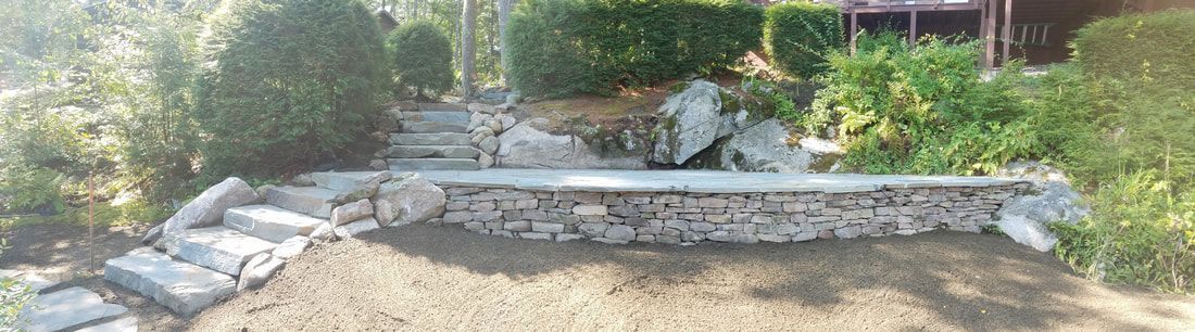 A stone retaining wall with matching stone steps in a landscaped garden with shrubs and trees.