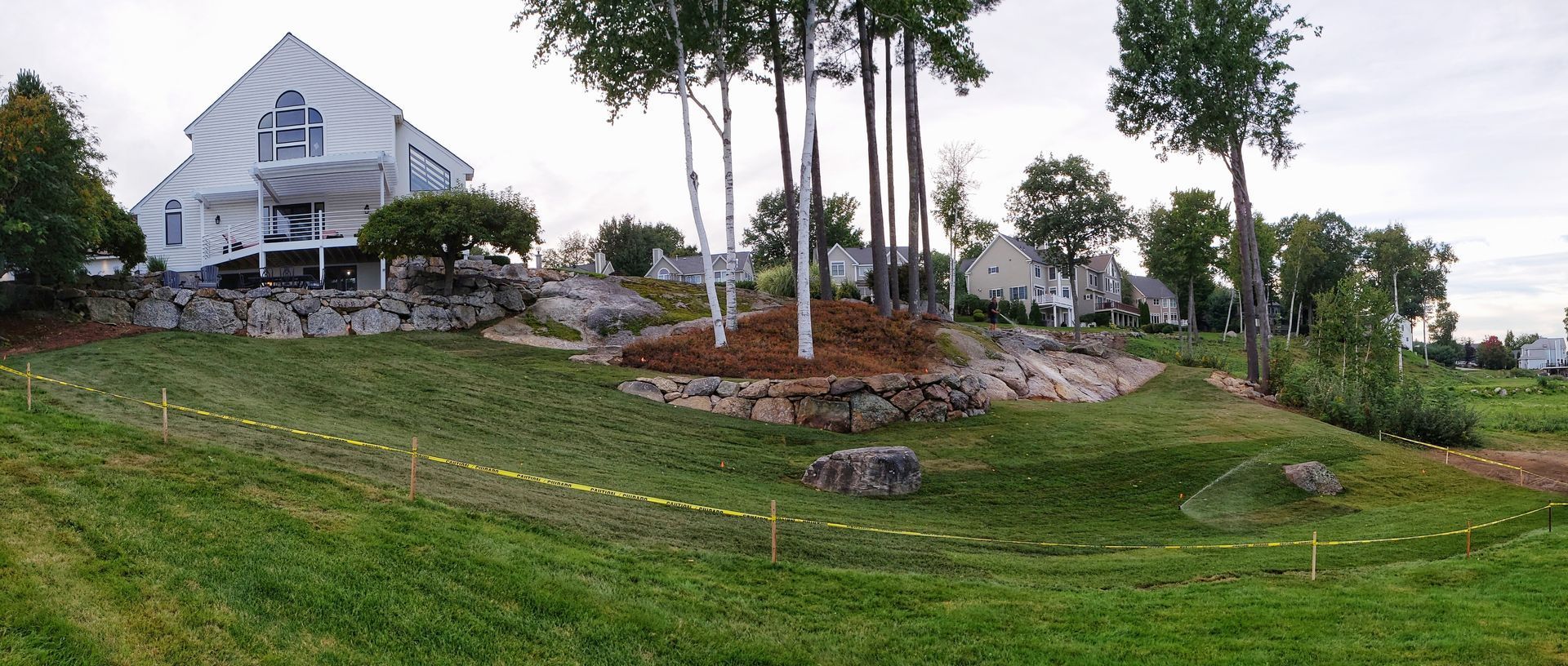 A wide panoramic view of a white multi-level house with stone retaining walls, surrounded by trees and grassy hills.