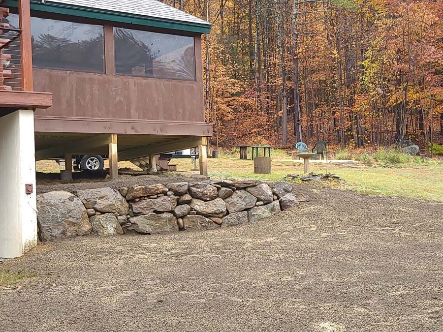 A rustic cabin porch supported by wooden posts stands behind a natural stone retaining wall in a wooded autumn setting.