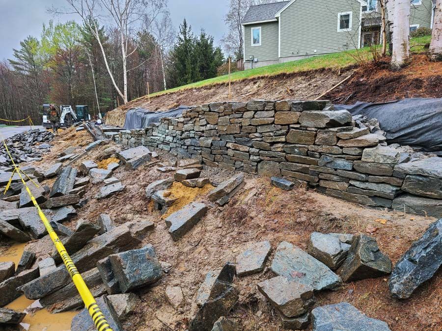 A stone retaining wall under construction on a slope with scattered rocks and construction tape in the foreground.