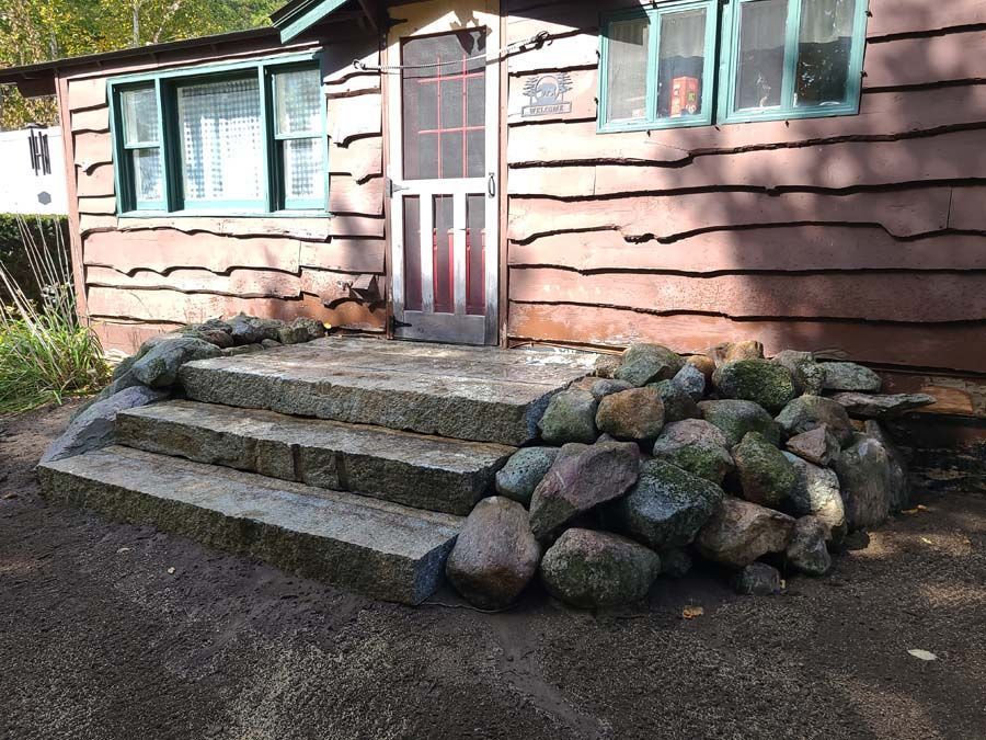 Stone steps leading to a rustic cabin entrance with brown siding, flanked by decorative rock landscaping.