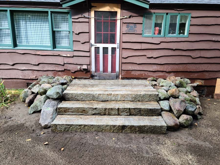 Three stone steps lead to a brown wooden cabin entrance, flanked by stacked decorative rocks on either side.