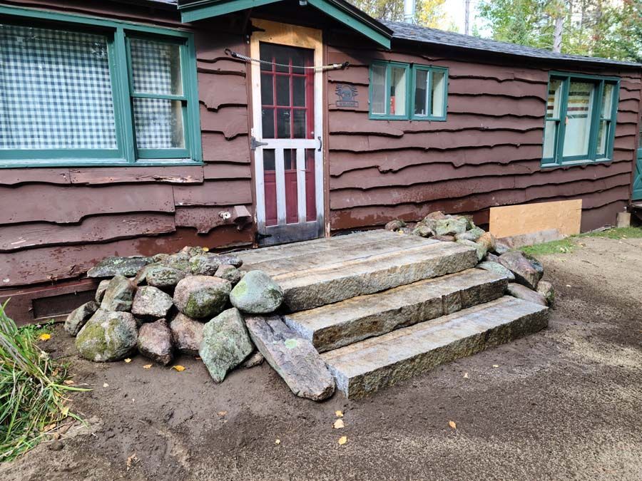 A cabin with dark brown horizontal siding, green window frames, and three stone steps leading to a screen door.
