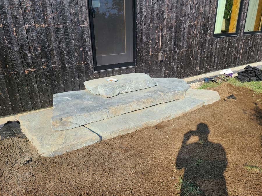 Stone steps leading to a dark wood-sided house entrance, with a shadow of a person visible in the foreground.