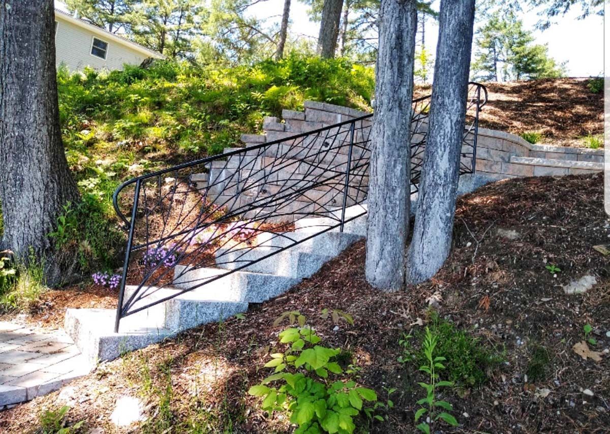 Stone steps ascend a gentle hill with a black, branch-patterned metal handrail, flanked by trees and greenery.