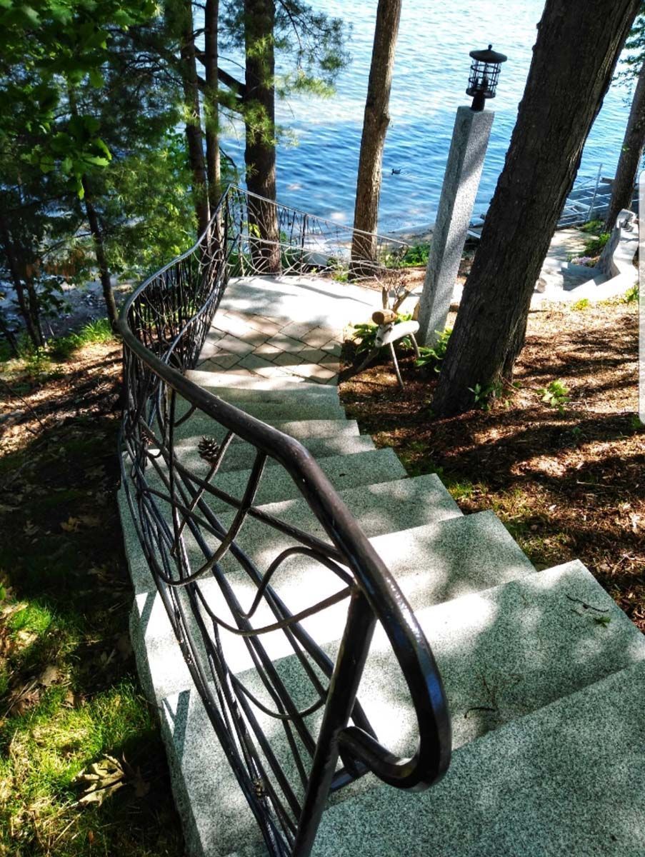 Stone steps with a decorative dark metal railing descend toward a lake surrounded by trees and a tall lamp post.