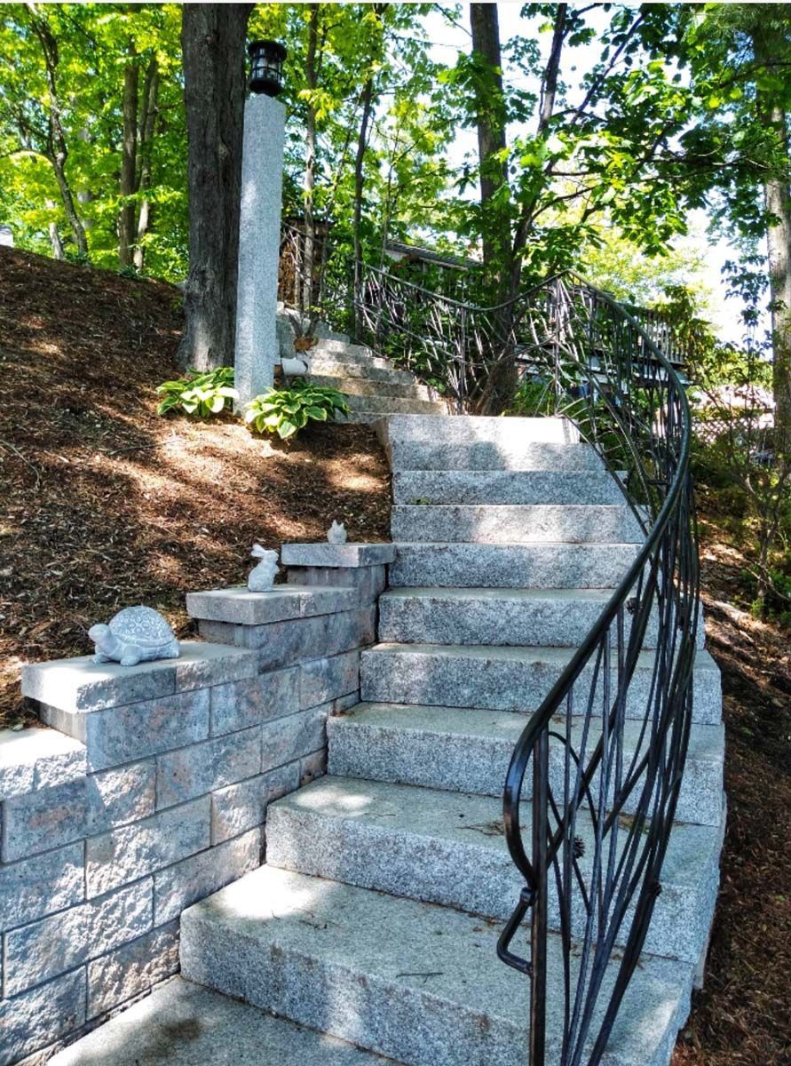 Outdoor stone steps with a black wrought iron railing, climbing a wooded hill next to a small stone retaining wall.