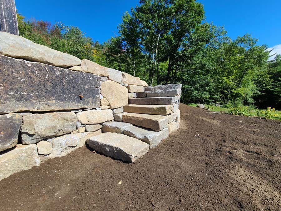 Stone steps built into a natural rock retaining wall outdoors next to a plot of dark, freshly raked soil.