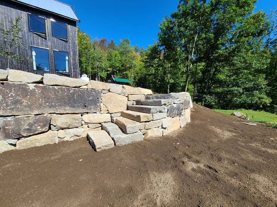 Stone steps built into a retaining wall next to a modern dark-paneled house and a sloping dirt yard.