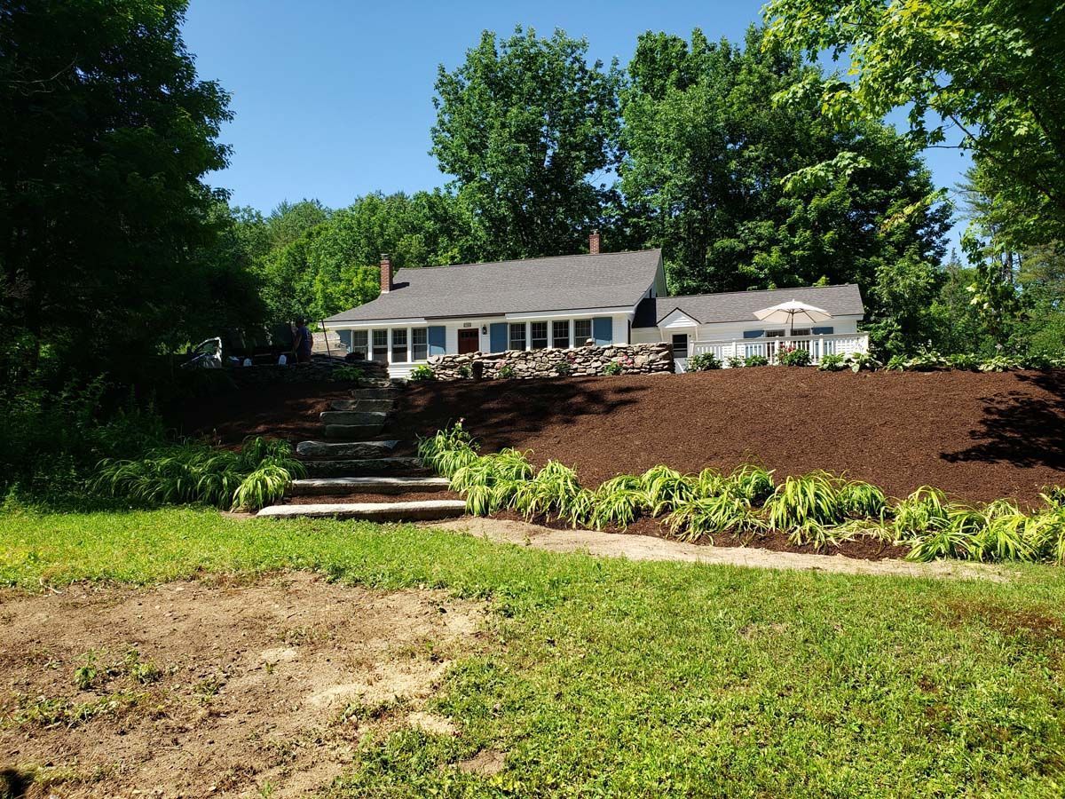 A single-story house with a grey roof sits on a hill above a landscaped yard with stone steps and mulch beds.