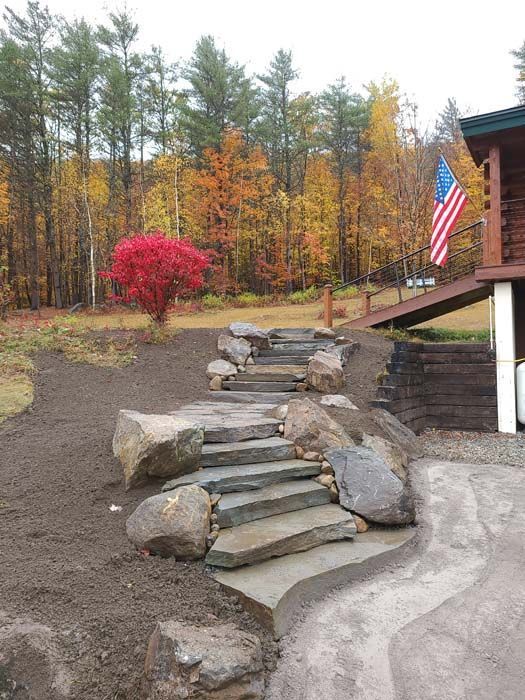 Stone steps lead up a hill toward a log house deck with an American flag, set against a backdrop of colorful autumn trees.