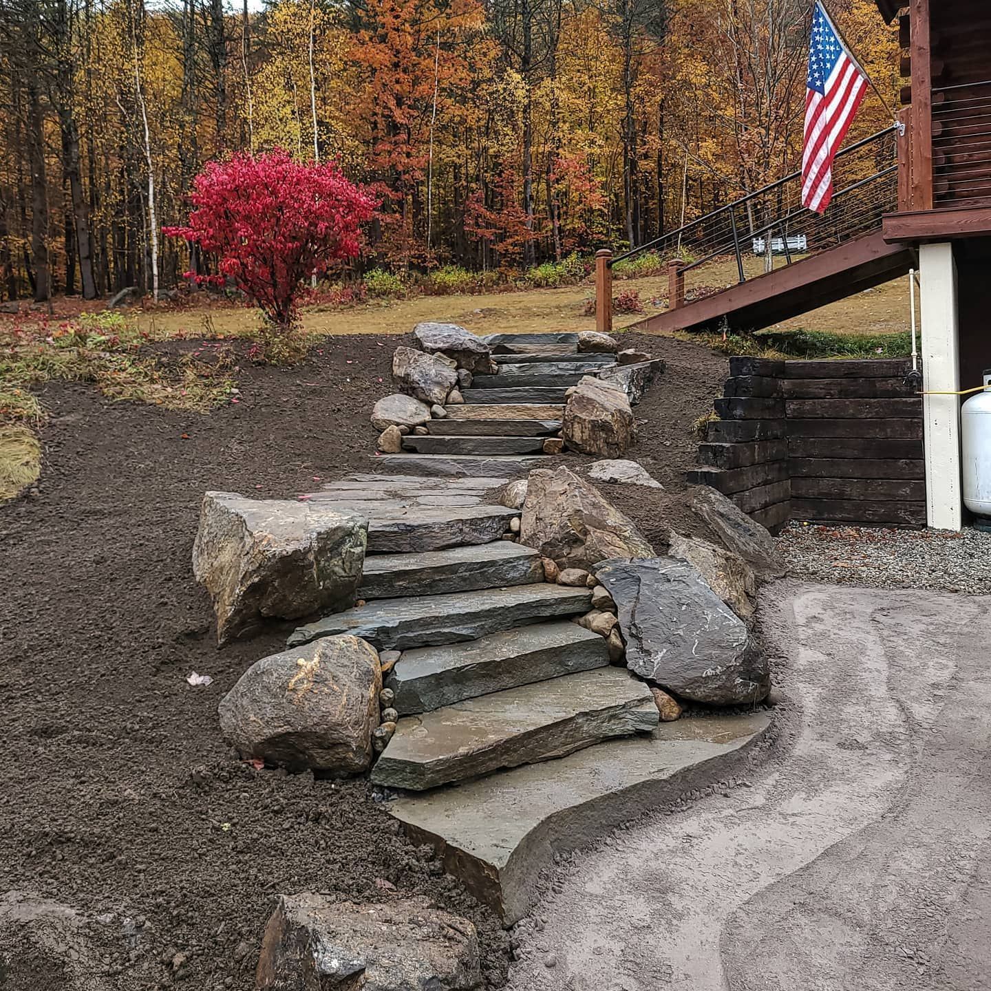 Stone steps lead up a hill toward a porch with an American flag, set against a background of autumn trees.