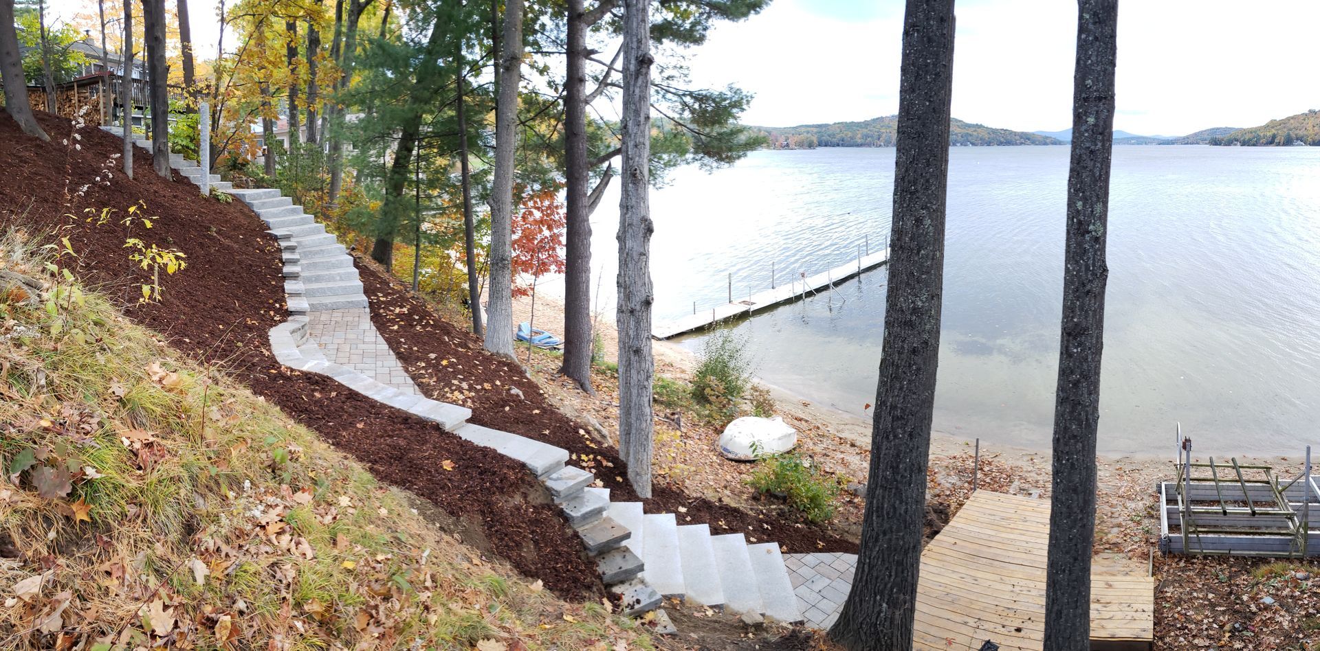 A stone staircase winds down a steep, wooded hillside to a lakeside dock and sandy beach on a clear day.