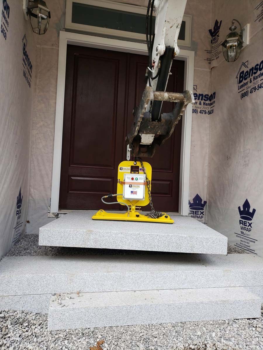 A machine uses a vacuum lifter to position a large, light gray stone slab for a porch step in front of a house entrance.