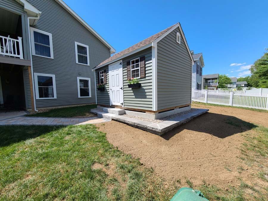 A grey storage shed sits on a stone patio next to a multi-story house under a bright blue sky.