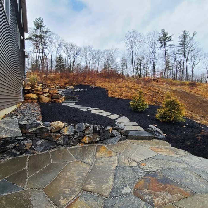 Flagstone patio transitions to a stone walkway winding through black mulch and evergreen shrubs beside a house exterior.
