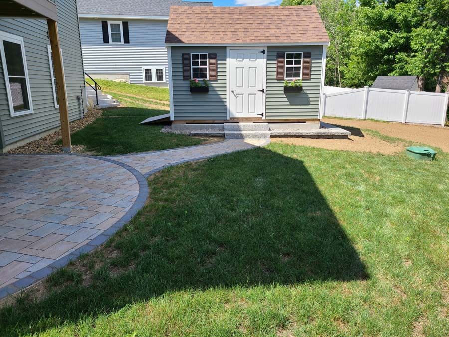 A sage green shed with a brown roof stands in a backyard, connected to a stone patio by a matching stone pathway.