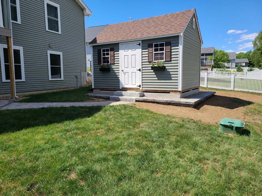 A gray shed with a white door and brown roof stands on a stone foundation in a backyard next to a matching house.