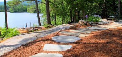 A stone stepping-stone path leads through a mulch-covered yard toward a lake with trees in the background.