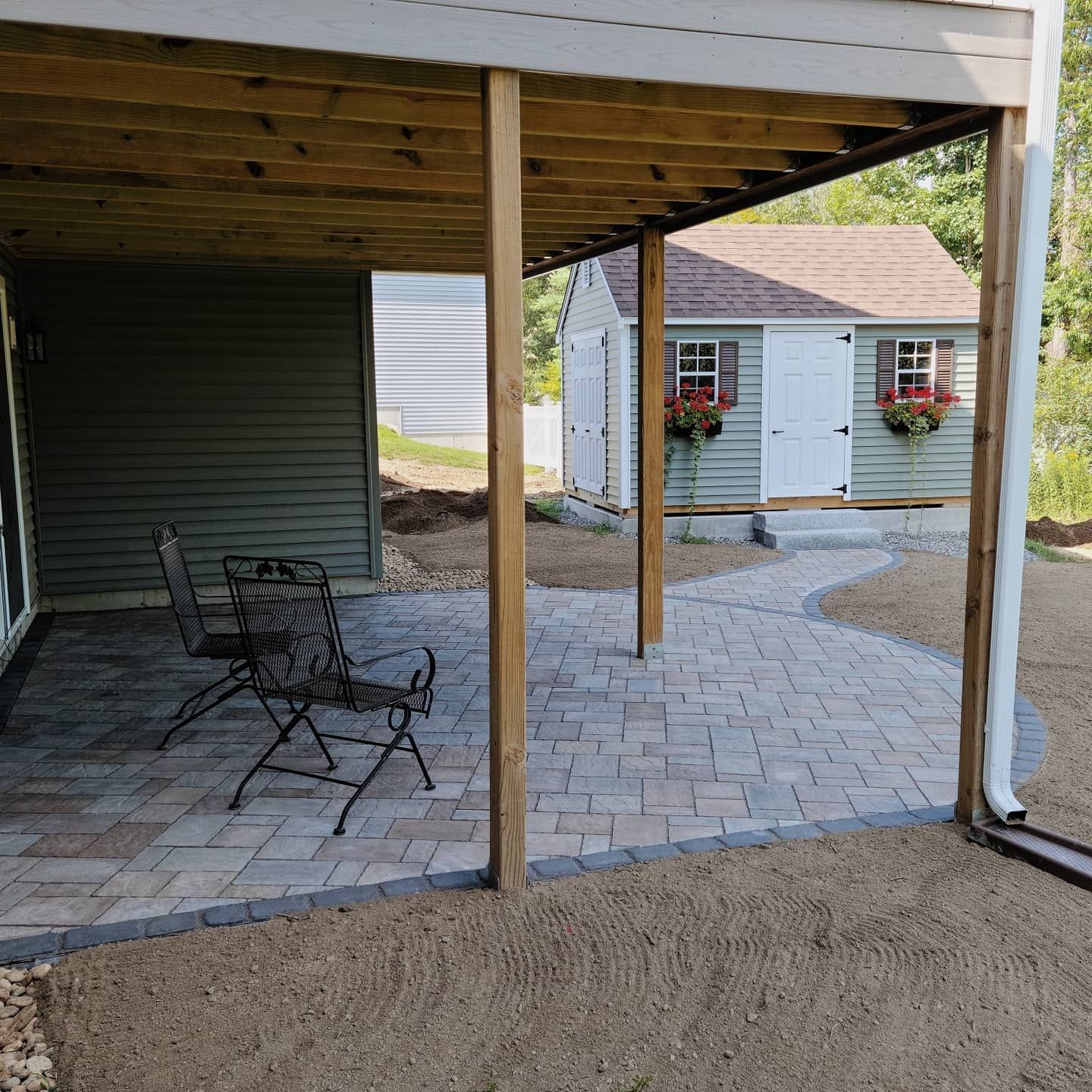 A paved stone patio under a deck, featuring two dark metal chairs, with a matching shed in the background.