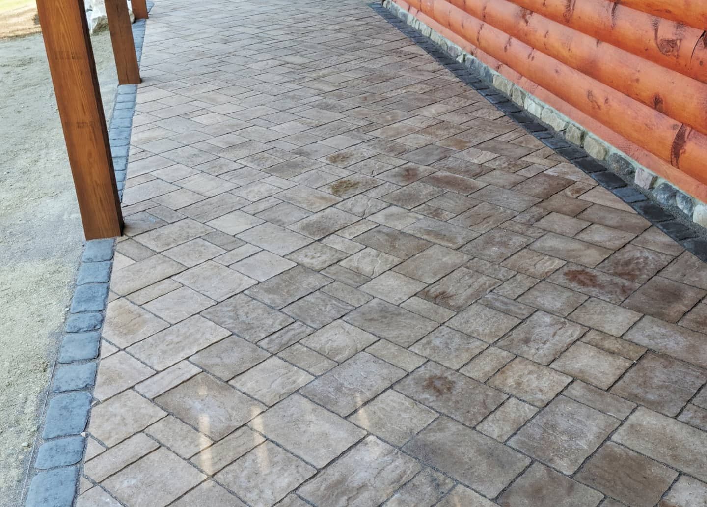 A patio paved with rectangular stone pavers in a herringbone pattern next to a log-style wall and wooden support posts.