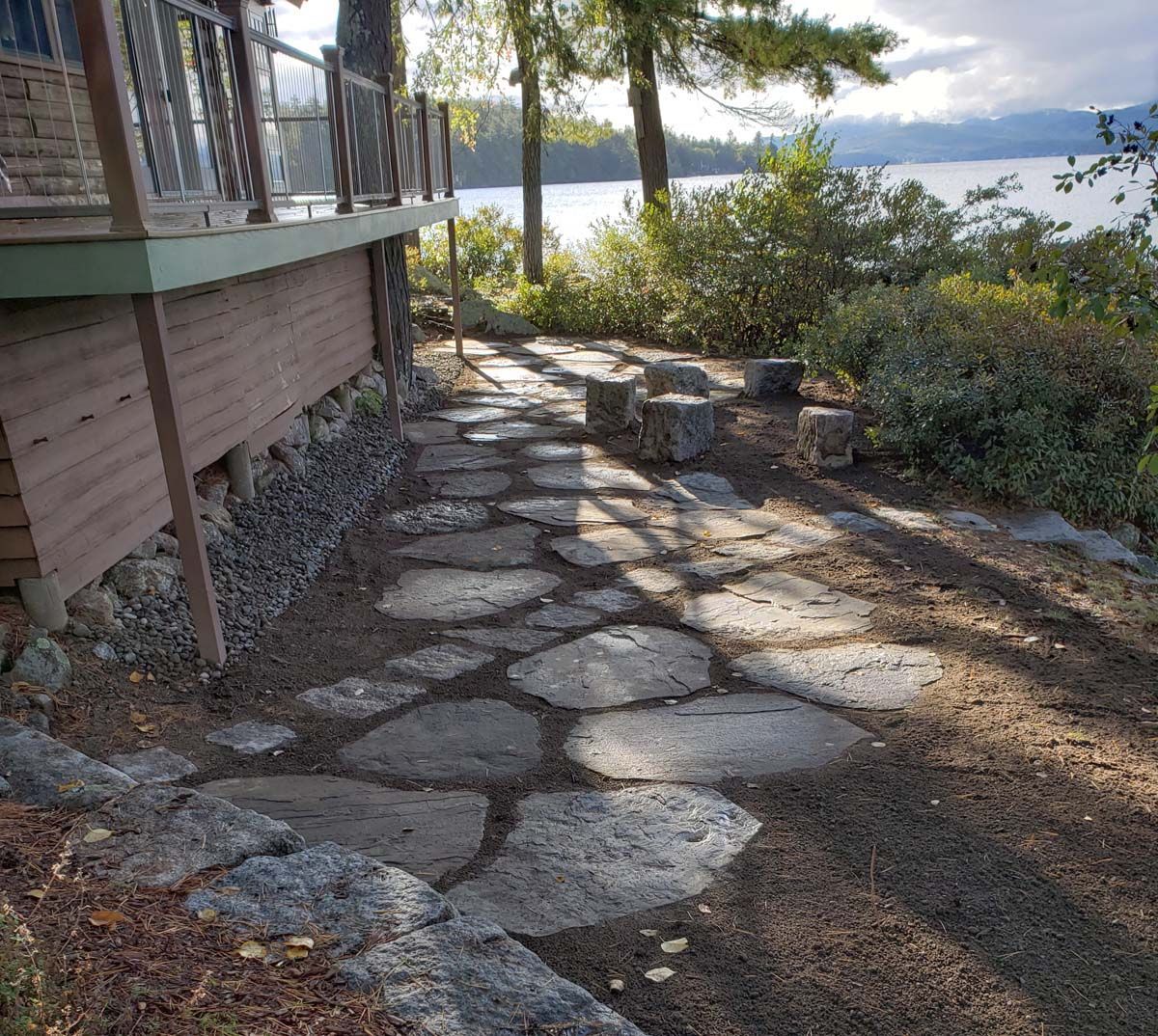 A flagstone walkway leads past a wooden deck toward a lake and trees under a cloudy sky.