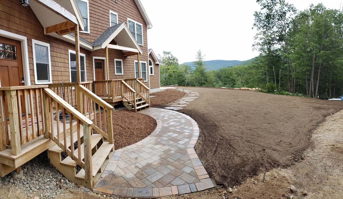 A new stone walkway leads to the wooden decks of a two-story home, bordered by landscaped beds and an expanse of soil.