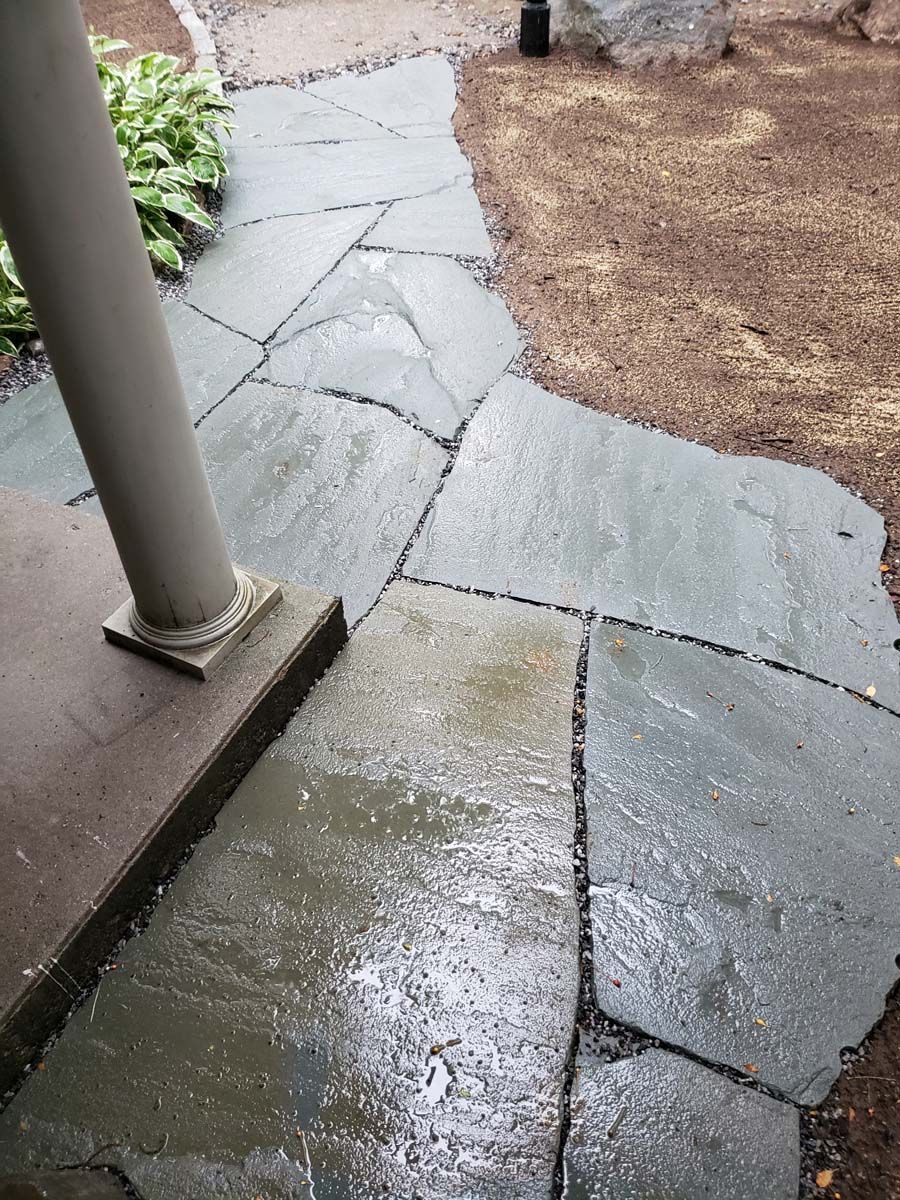 A wet, gray flagstone walkway leading toward a porch post next to a garden bed and mulched yard.