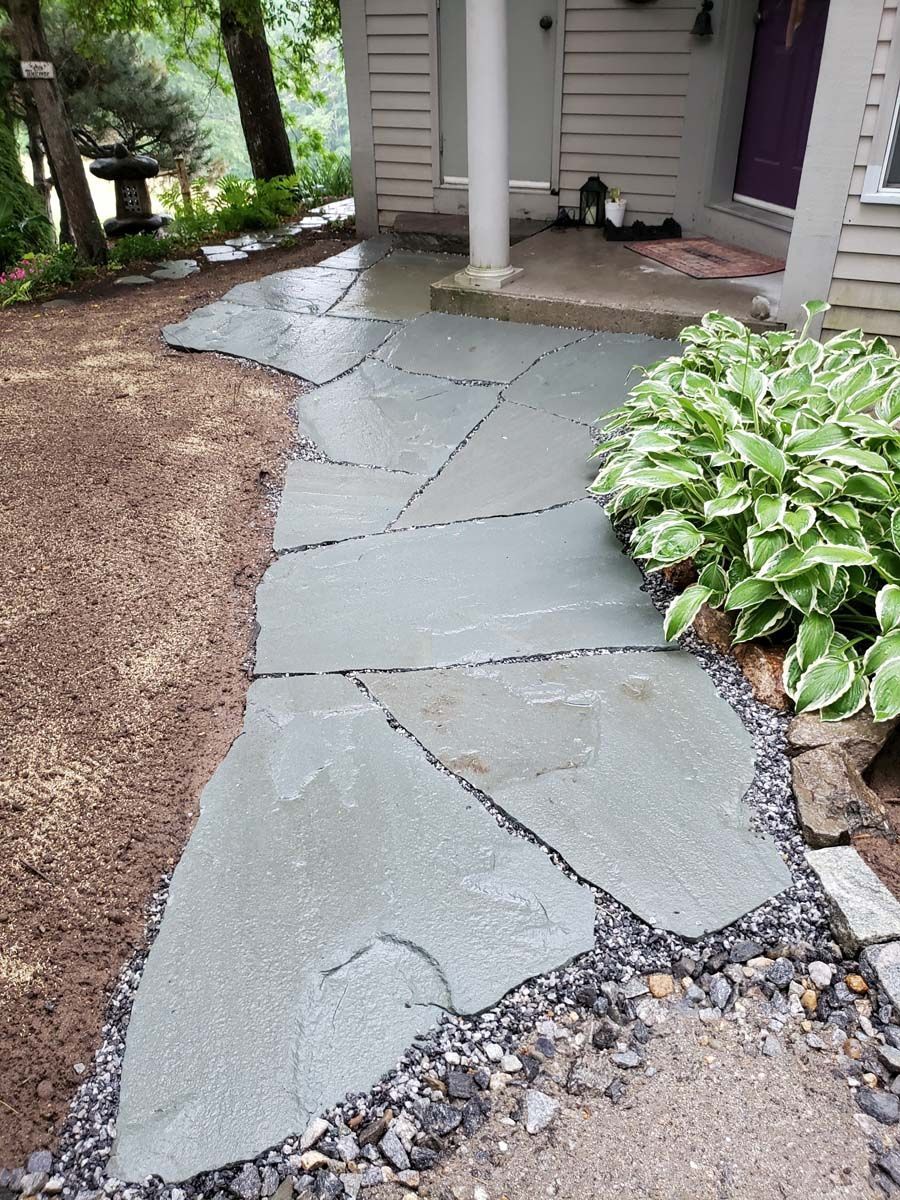 A flagstone walkway made of large, irregular grey stones leads to the front porch of a house with light-colored siding.