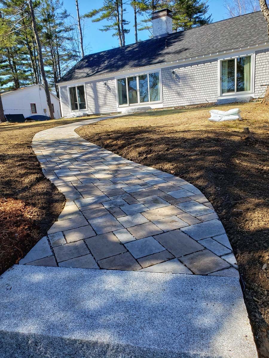 A light-colored stone walkway curves through a lawn toward a house on a sunny day.