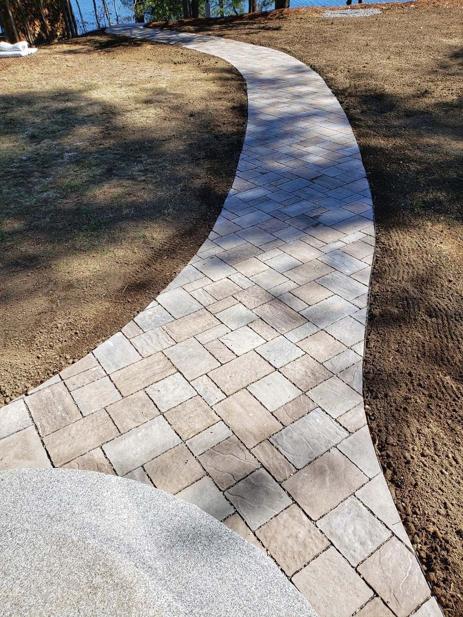 A curving stone paver walkway leads through a yard toward a body of water in the background.