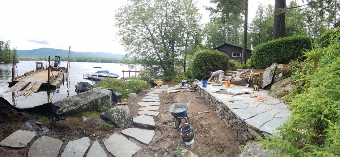A landscape construction site by a lake features a stone walkway and patio under construction near a wooden dock.
