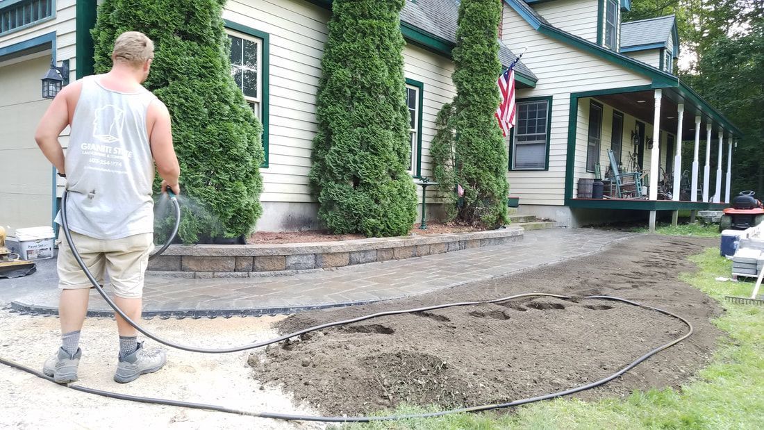 A person installs landscape edging along a stone pathway in front of a yellow house with a covered porch.