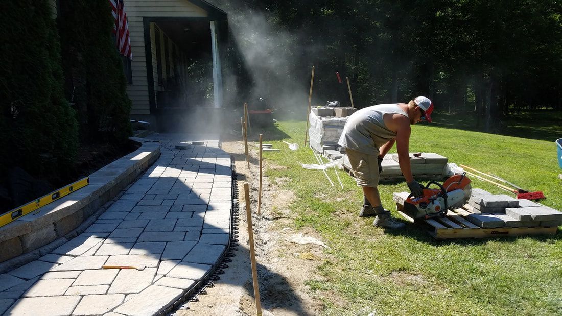 A person in a gray tank top and shorts cuts stone pavers with a gas-powered saw on a sunny residential lawn.
