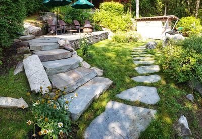 A stone staircase leads to an upper patio with patio furniture and umbrellas, next to a flagstone path through a garden.