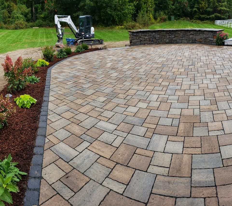 A wide, curved stone paver patio sits next to a landscaped garden bed, with a small excavator parked on the lawn behind it.