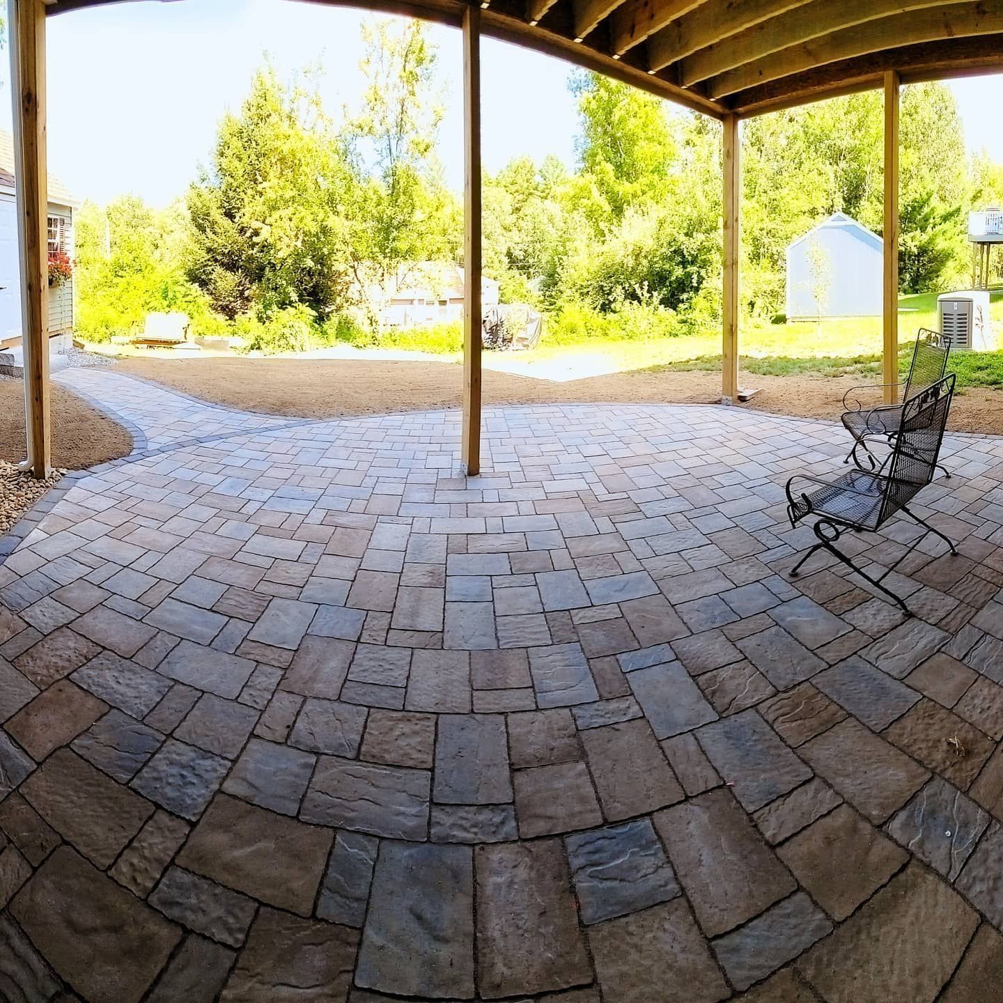 A stone patio with a circular design pattern under a wooden deck, with a metal chair and a sunny backyard in the distance.