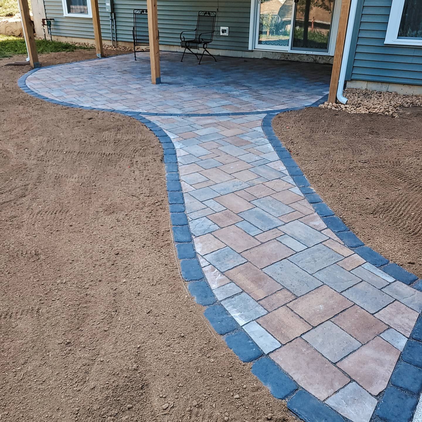 A newly installed paver walkway with a dark border leads to a circular patio in front of a house with blue siding.