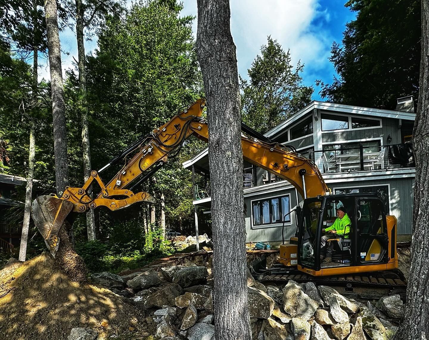A yellow excavator operated by a person in a high-visibility jacket works on a rocky landscape in front of a modern house.