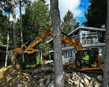A yellow excavator operated by a person in a high-visibility jacket works on a rocky landscape in front of a modern house.