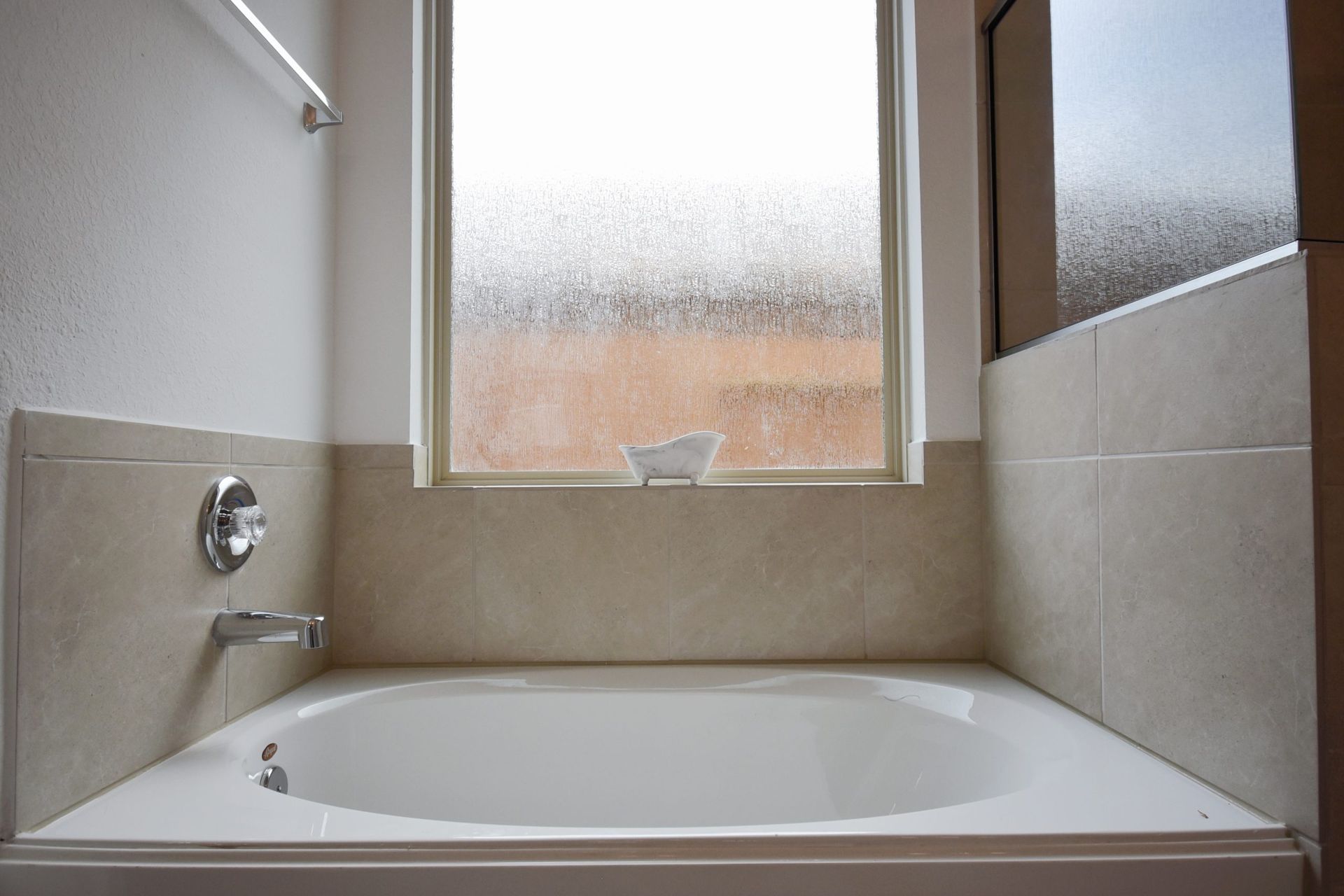 Oval bathtub beneath a window in a bathroom. Beige tiles surround the tub and window.